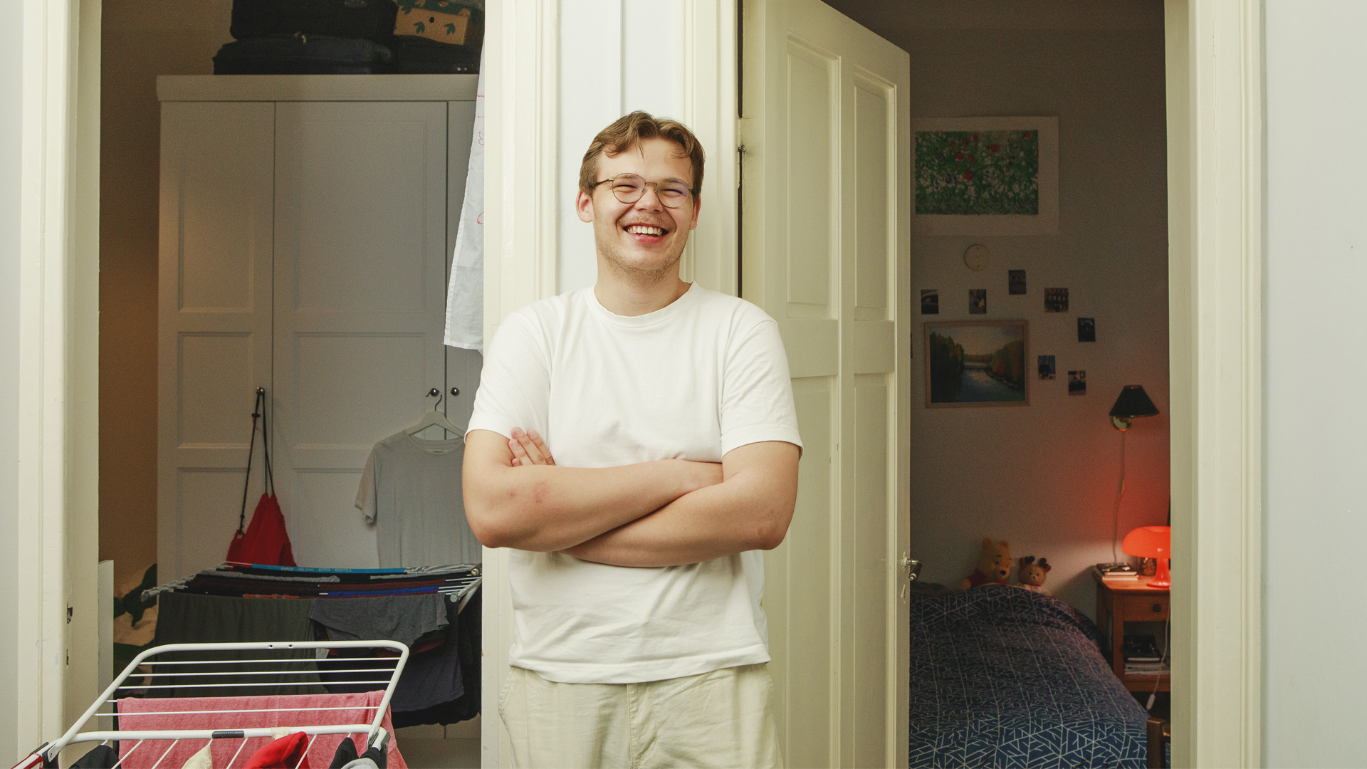 Nuori valkoinen mies nojaa seinään valkoisessa asussa hymyillen, takana kaksi oviaukkoa, joista näkyy kaksi makuuhuonetta.